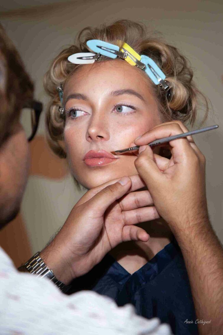 A business photograph depicting a model's face receiving hairstyling and makeup application by an expert hand, with the makeup artist's face subtly blurred in the foreground, illustrating the artistry of professional beauty services.
