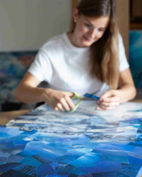 An image showcasing a collage artist's canvas with a vivid blue sky taking focus, while the artist works in the blurry background, symbolizing the harmony of art and nature, and the artist's humble role in her creation.