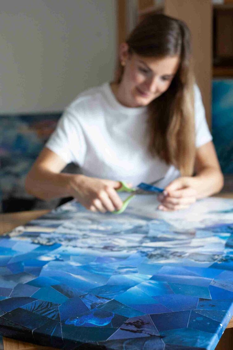 An image showcasing a collage artist's canvas with a vivid blue sky taking focus, while the artist works in the blurry background, symbolizing the harmony of art and nature, and the artist's humble role in her creation.